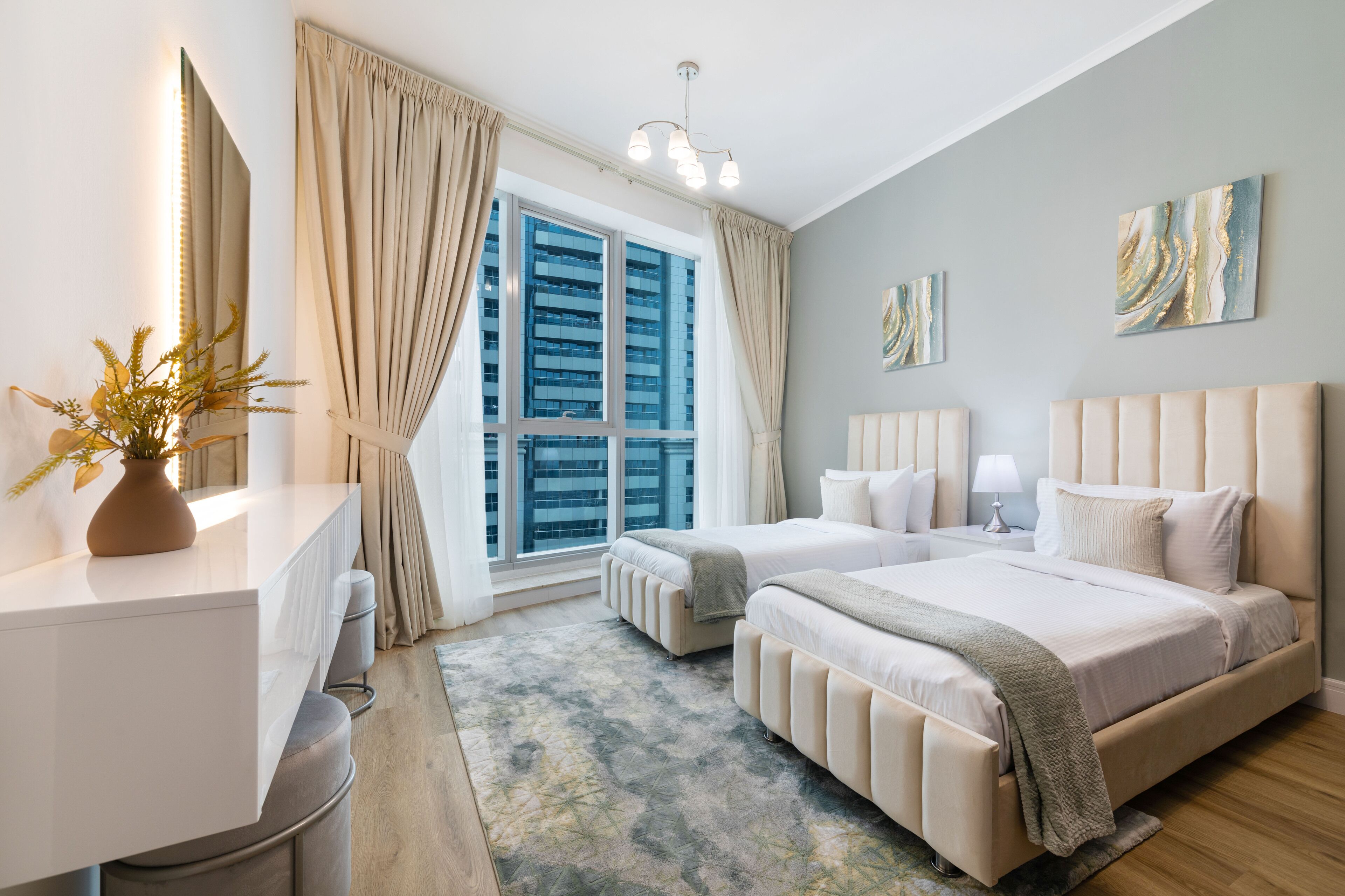 Ein modernes Hotelzimmer mit zwei Betten, einem großen Fenster mit Vorhängen, einem Nachttisch mit Lampe und Kunstwerken an den Wänden.