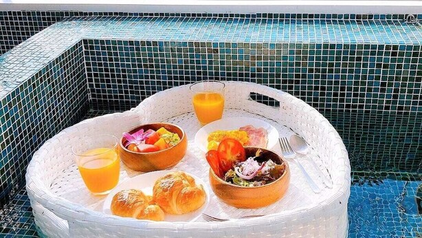 Desayuno con cruasanes, fruta y un vaso de zumo de naranja sobre una mesa de mimbre blanco junto a una piscina.