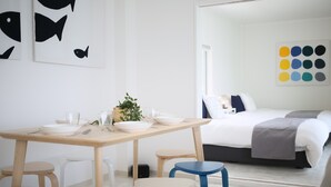 Una habitación de hotel moderna y minimalista con una mesa de comedor de madera, taburetes blancos, una cama con una manta gris y una pared con arte abstracto.
