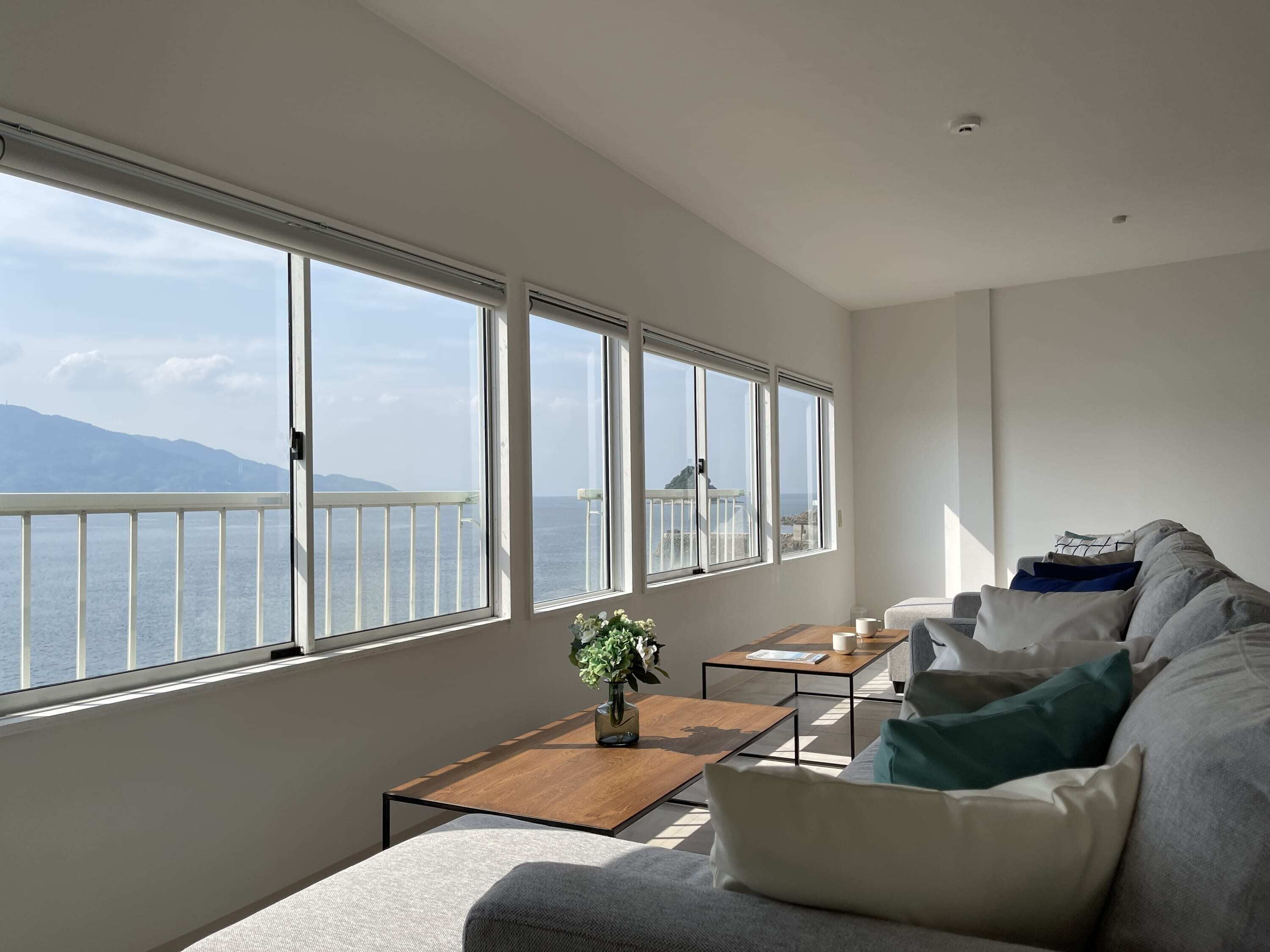 A modern living room with a large window overlooking a balcony and the sea. There is a sofa, a coffee table, and a small side table with a plant.