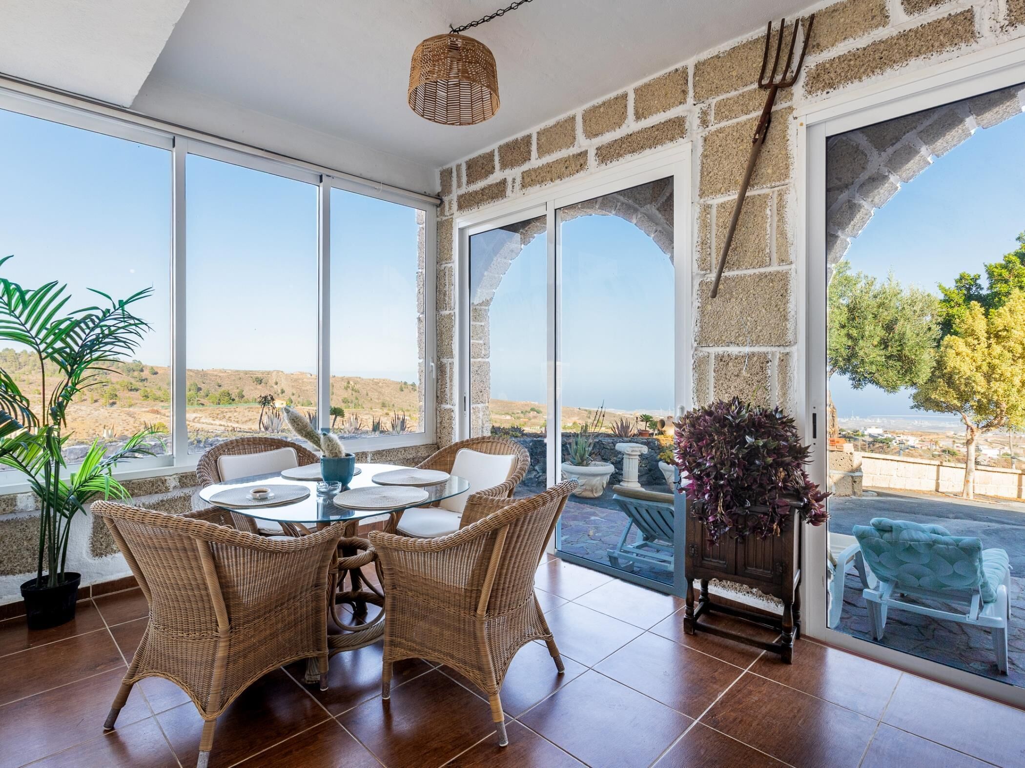 A dining area with wicker furniture, a glass table, and a view of the landscape through large windows.