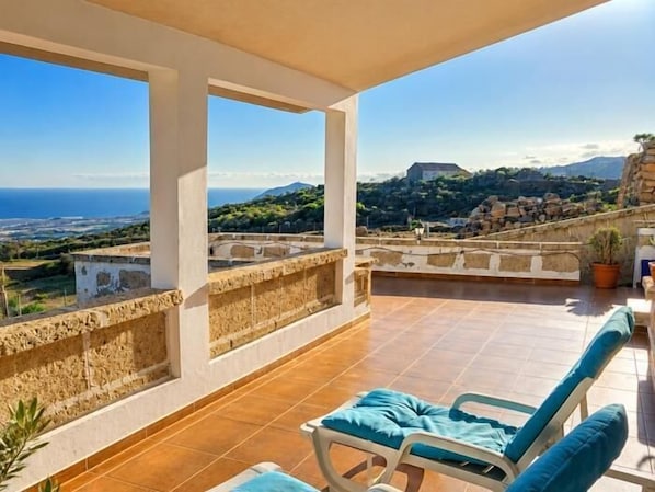A rooftop terrace with a view of the sea and hills, a lounge chair, and a potted plant.