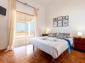 Une chambre à coucher comprenant un lit, une table de chevet, une lampe, un mur orné de tableaux encadrés et une porte-fenêtre coulissante donnant sur un balcon.