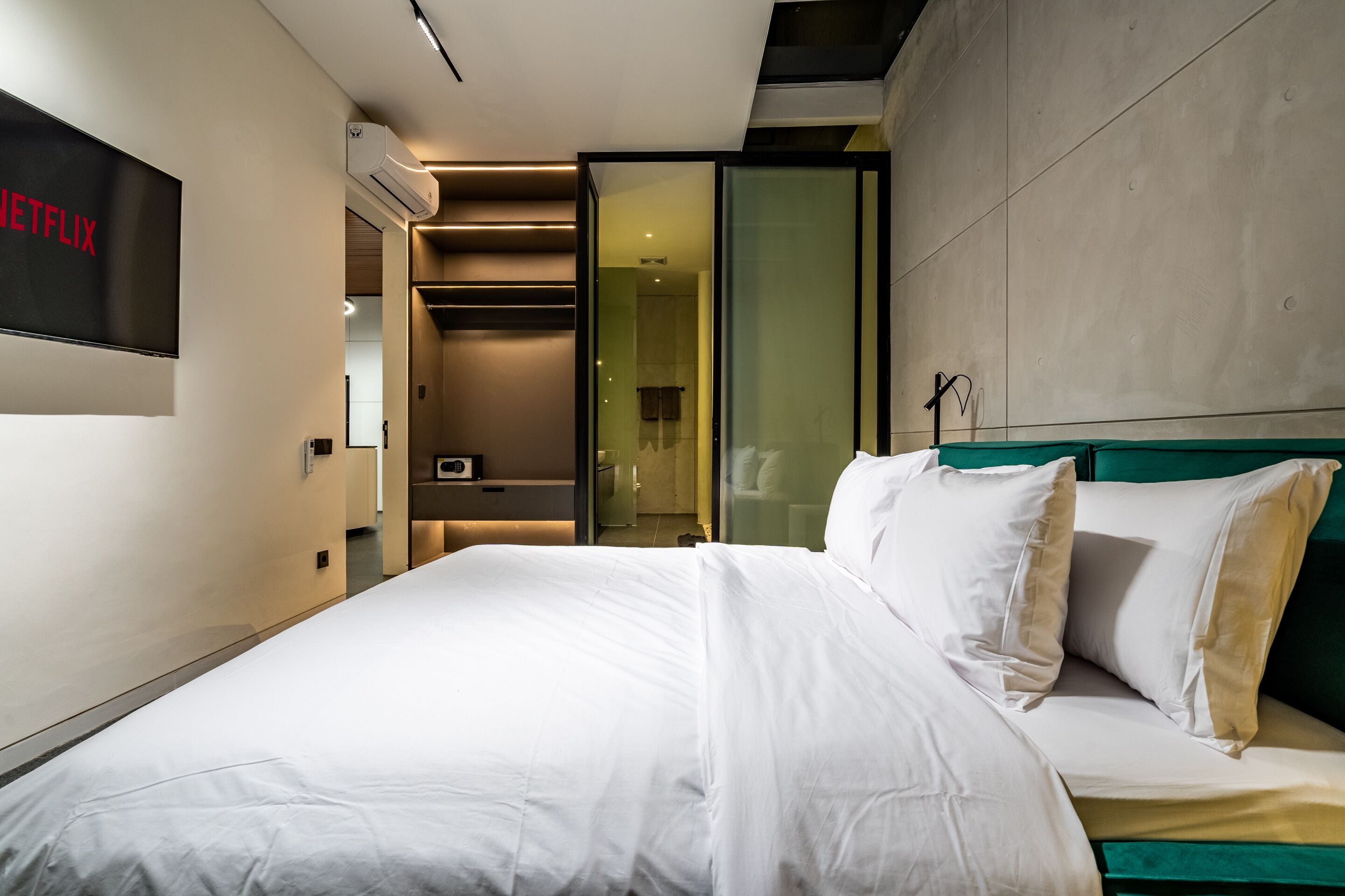 A modern hotel room with a large bed, a TV screen displaying the Netflix logo, and a glass door leading to a bathroom.