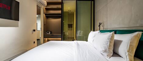 A modern hotel room with a large bed, a TV screen displaying the Netflix logo, and a glass door leading to a bathroom.