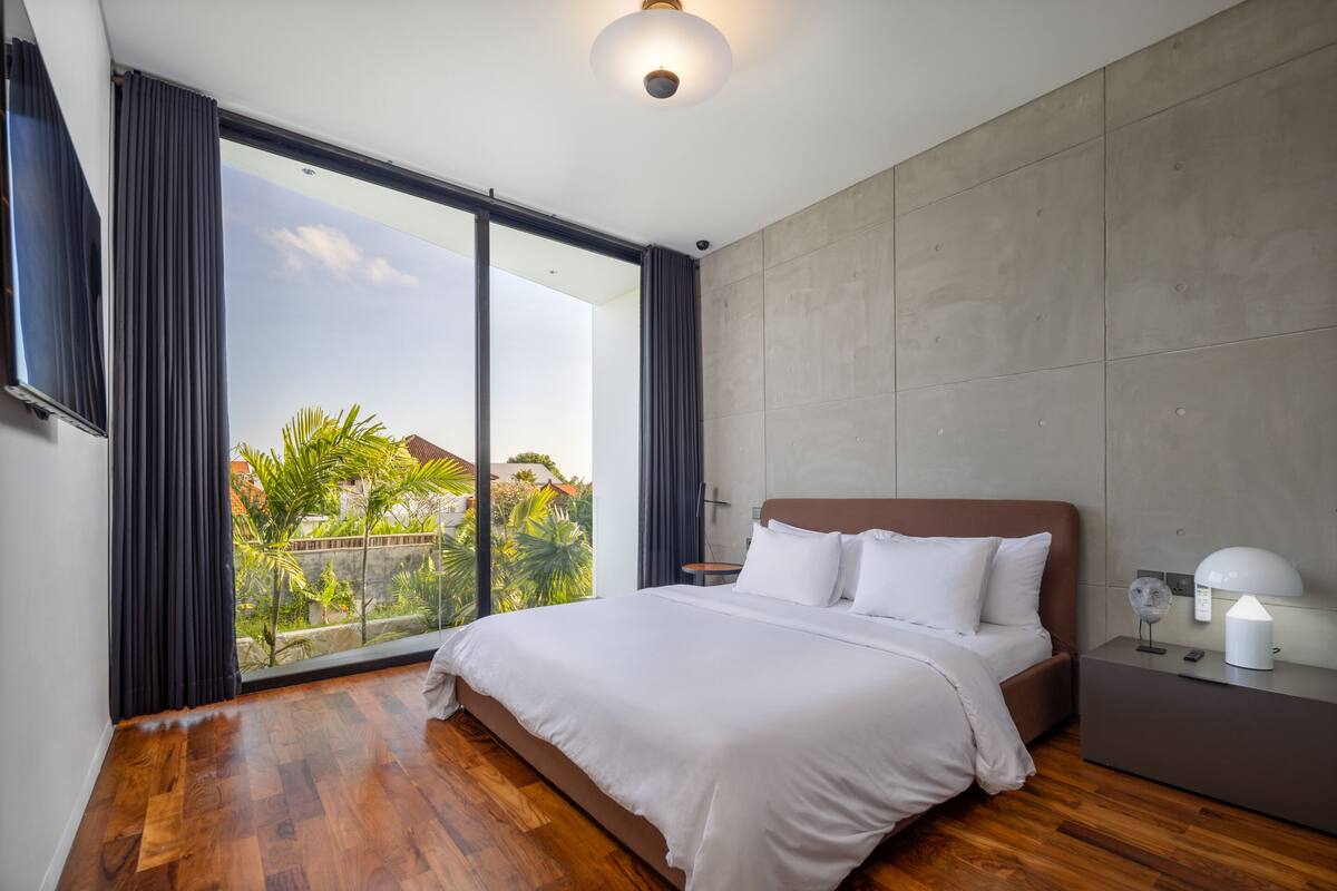 A modern bedroom with a large bed, a nightstand, and a view of greenery through floor-to-ceiling windows.