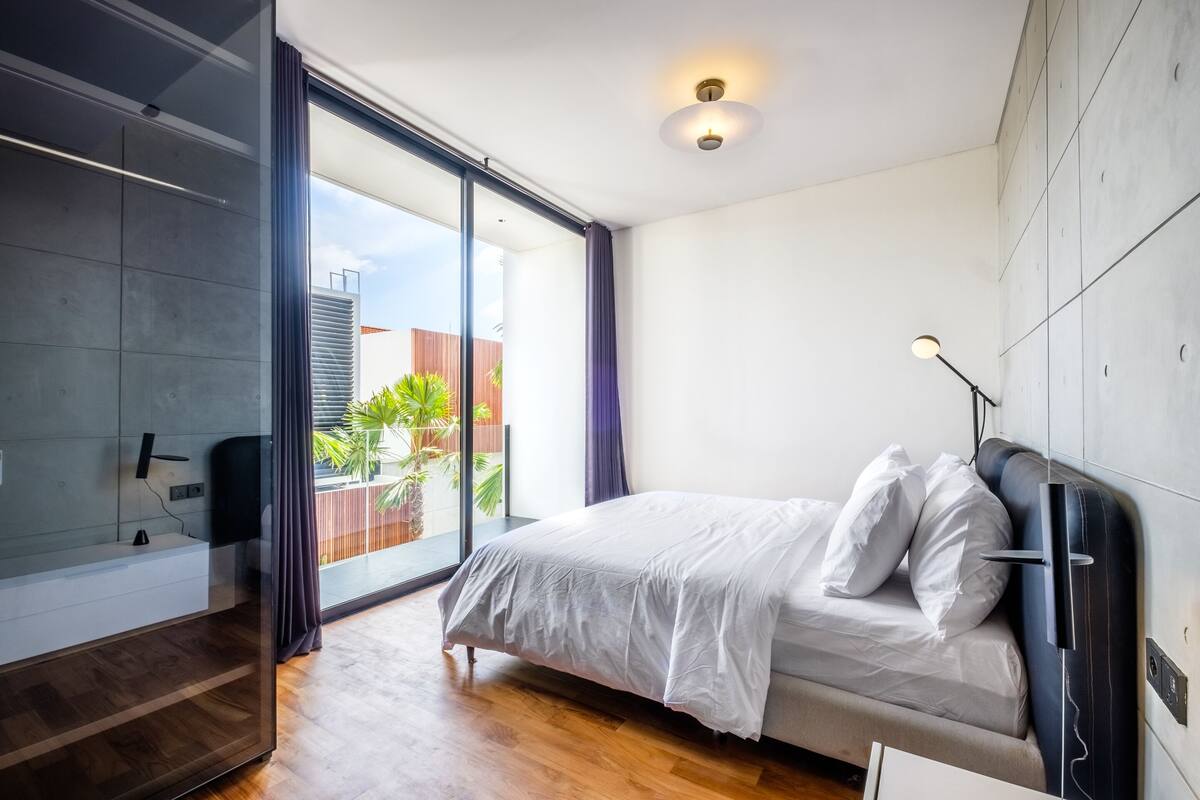 A modern bedroom with a large bed, a built-in TV, and a sliding glass door leading to a balcony.