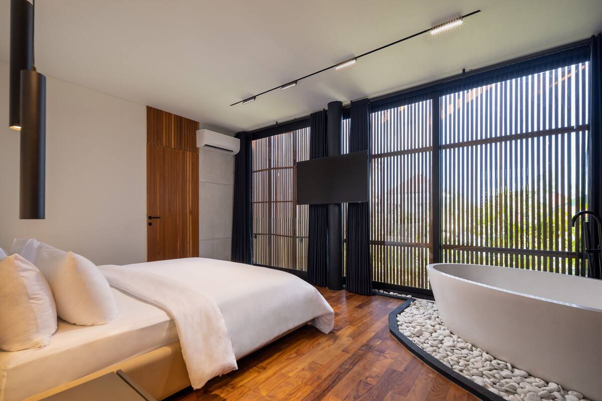A modern bedroom with a large bed, a bathtub, and a view through bamboo blinds.