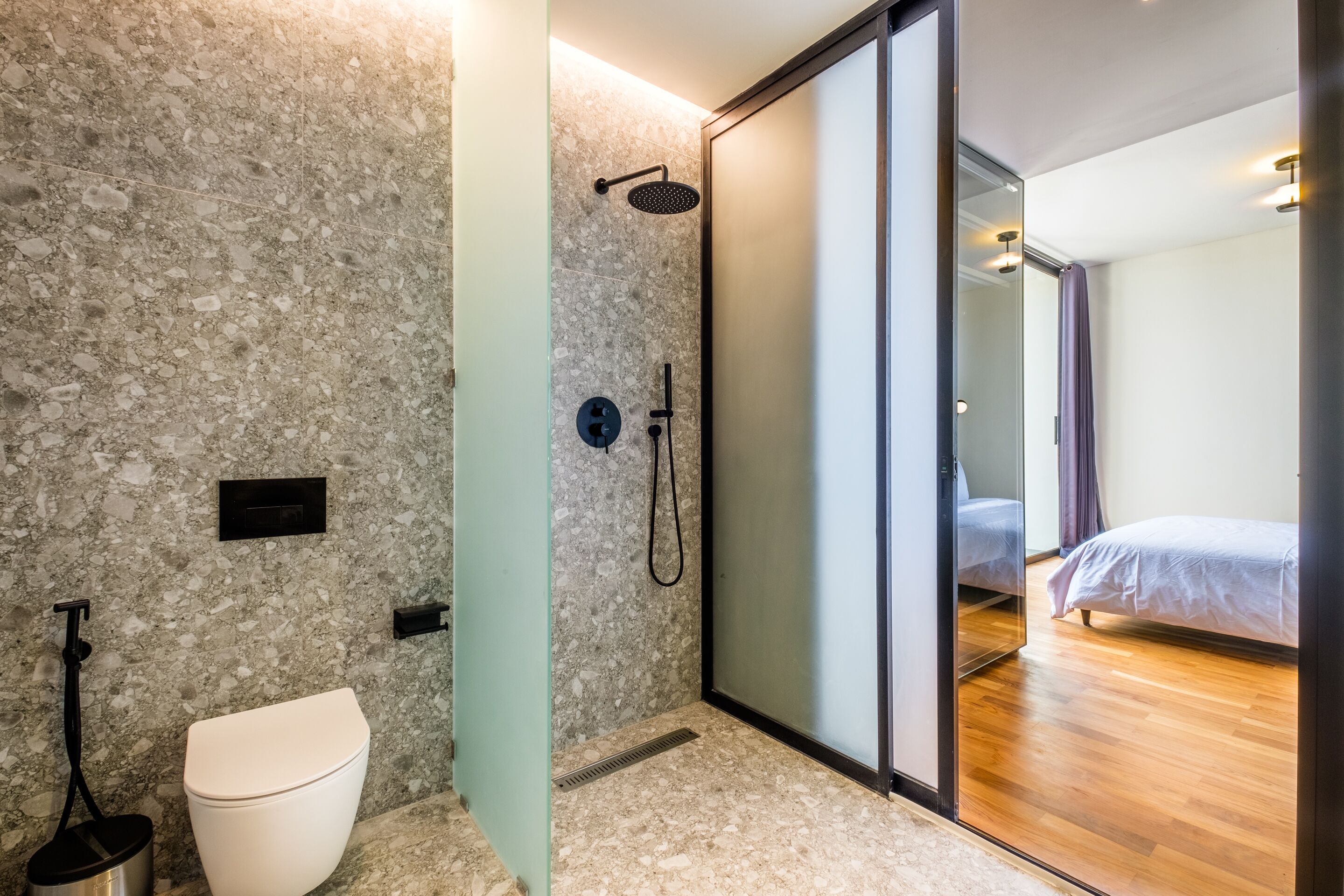 Un baño moderno con ducha, inodoro y una puerta de cristal que da a un dormitorio con cama.