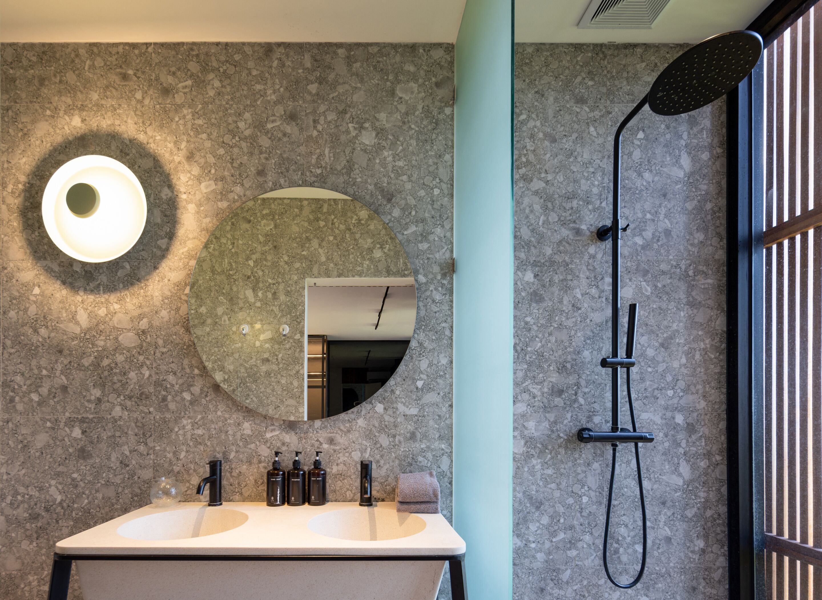 A bathroom with a marble wall, a round mirror, a sink with two basins, and a shower with a black hose.