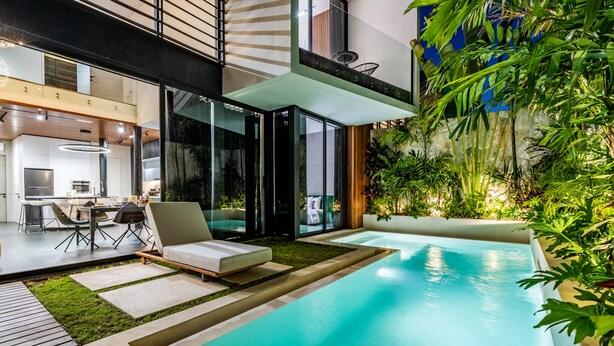 A modern outdoor pool area with a lounge chair, a dining table with chairs, and a kitchen visible through glass doors. - Alex Villas Complex N1 (Canggu)