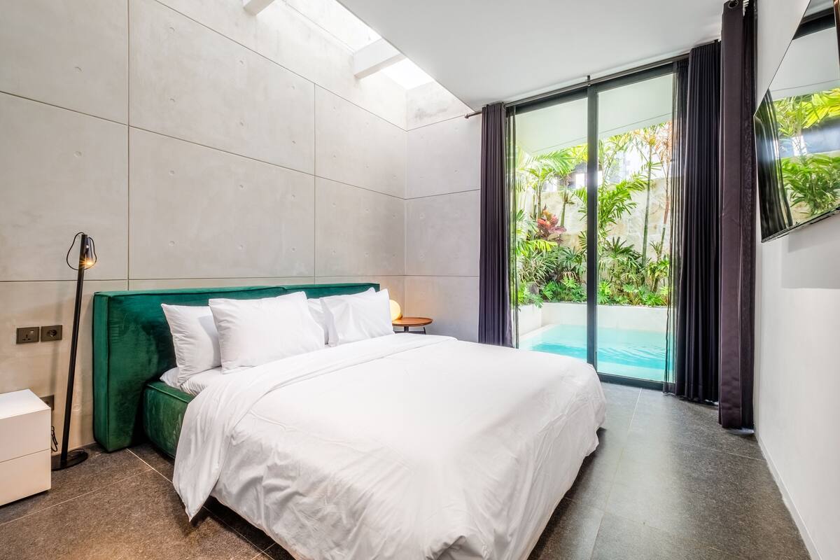 A modern hotel room with a large bed, a nightstand, a lamp, and a view of a swimming pool through a glass door.
