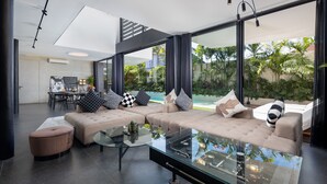 A modern living room with a large sectional sofa, a glass coffee table, and a view of a swimming pool and greenery outside. - Alex Villas Complex N1 (Canggu)