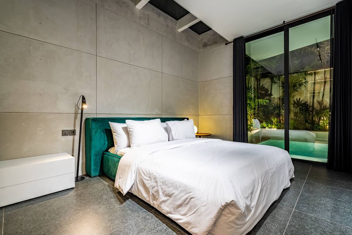 A modern bedroom with a large bed, a built-in bench, and a glass door leading to a balcony with greenery.