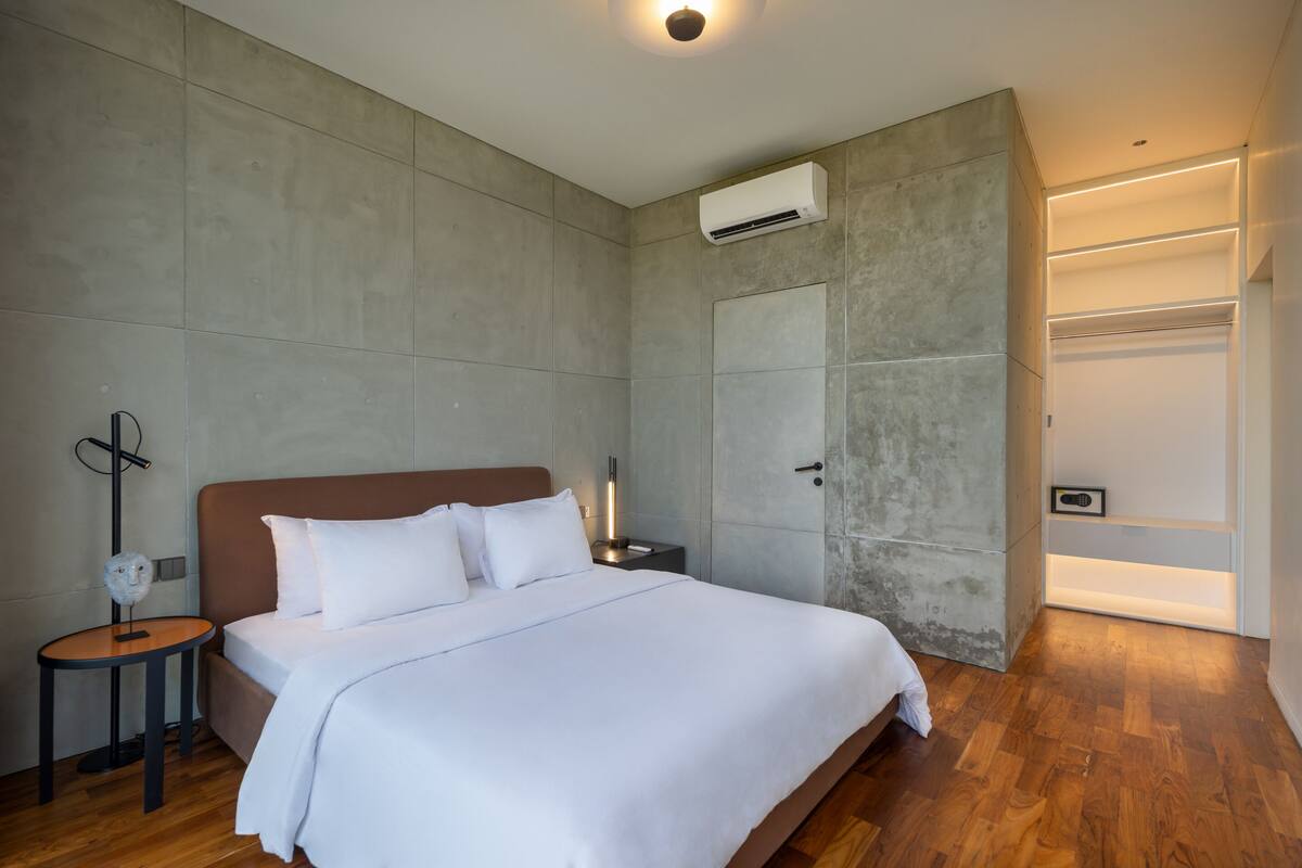 A modern hotel room with a large bed, a bedside table, an air conditioning unit, and a wardrobe.