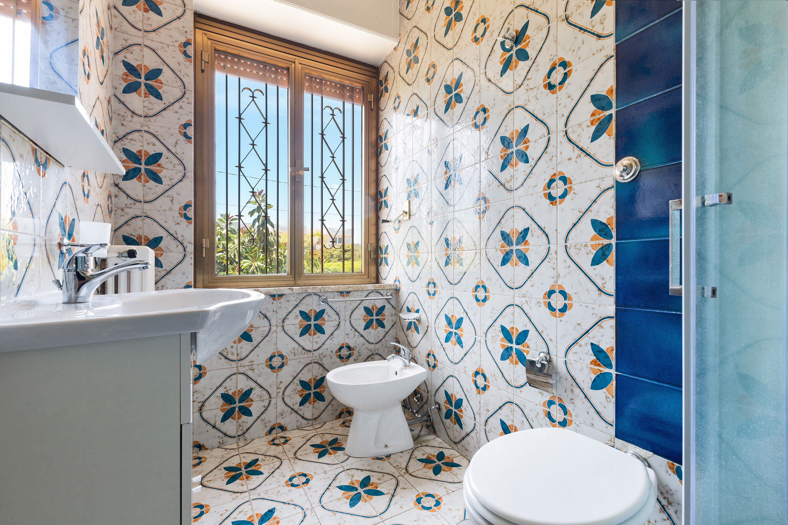 Casa de banho com lavatório branco, lavatório sobreposta branco e sanita branca, com janela e grades de metal.