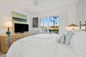 A bedroom with a bed, a TV, a ceiling fan, a window with a view of palm trees, and a painting on the wall. - Hidden Gem in Palm Desert, Steps to Living Desert! (Palm Desert)