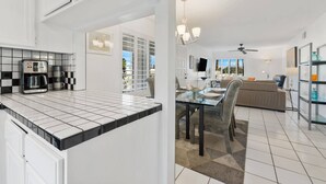 Eine moderne Küche mit Kaffeemaschine, ein Essbereich mit Glastisch und Stühlen sowie ein Wohnzimmer mit Sofa und Fernseher.