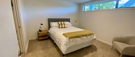A bedroom with a bed, bedside table, lamp, chair, and a window with a view of trees.