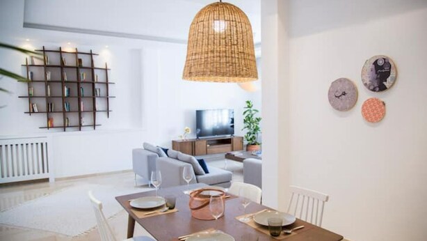A modern dining area with a wooden table set for four, a hanging woven light fixture, and a bookshelf in the background.