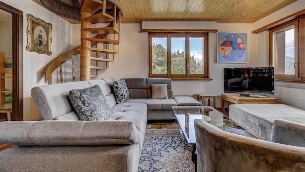 A modern living room with a curved wooden staircase, a large sectional sofa, a television, and a view of the outdoors.
