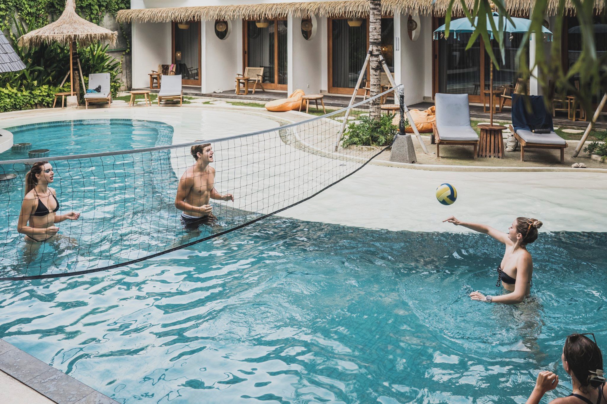 A pool area with a volleyball net, lounge chairs, and a thatched-roof structure.