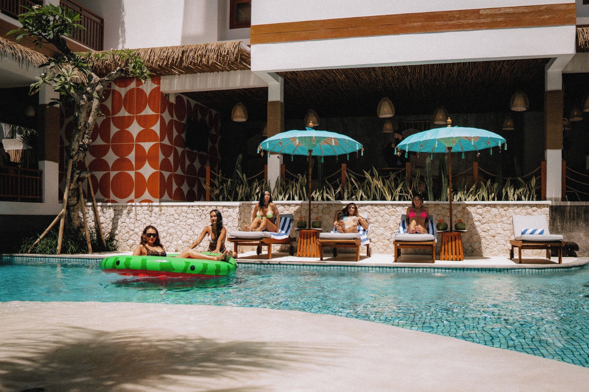 A pool area with lounge chairs and umbrellas, surrounded by a stone wall and tropical plants.
