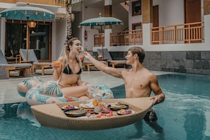 Una pareja en una piscina con una mesa flotante para comer, rodeada de sillones y sombrillas.