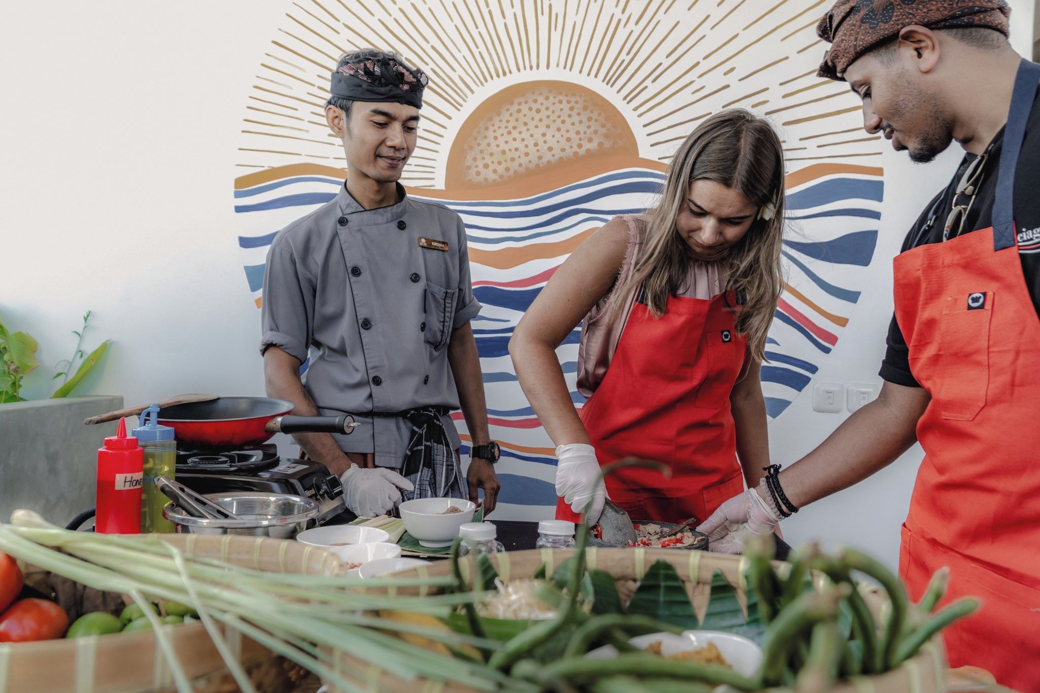 Three individuals in a kitchen setting with cooking utensils and ingredients, a patterned wall in the background.