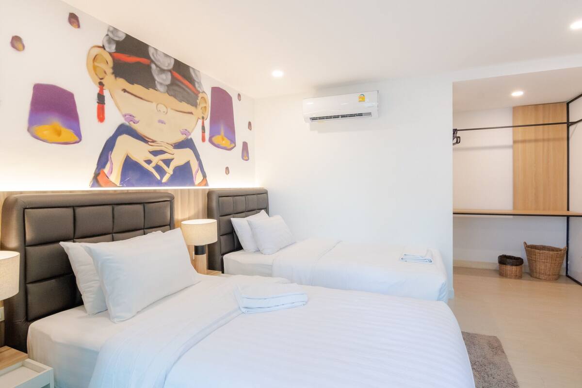 Ein Hotelzimmer mit zwei Betten, einem großen Wandbild, einer Klimaanlage und einem Holzregal.