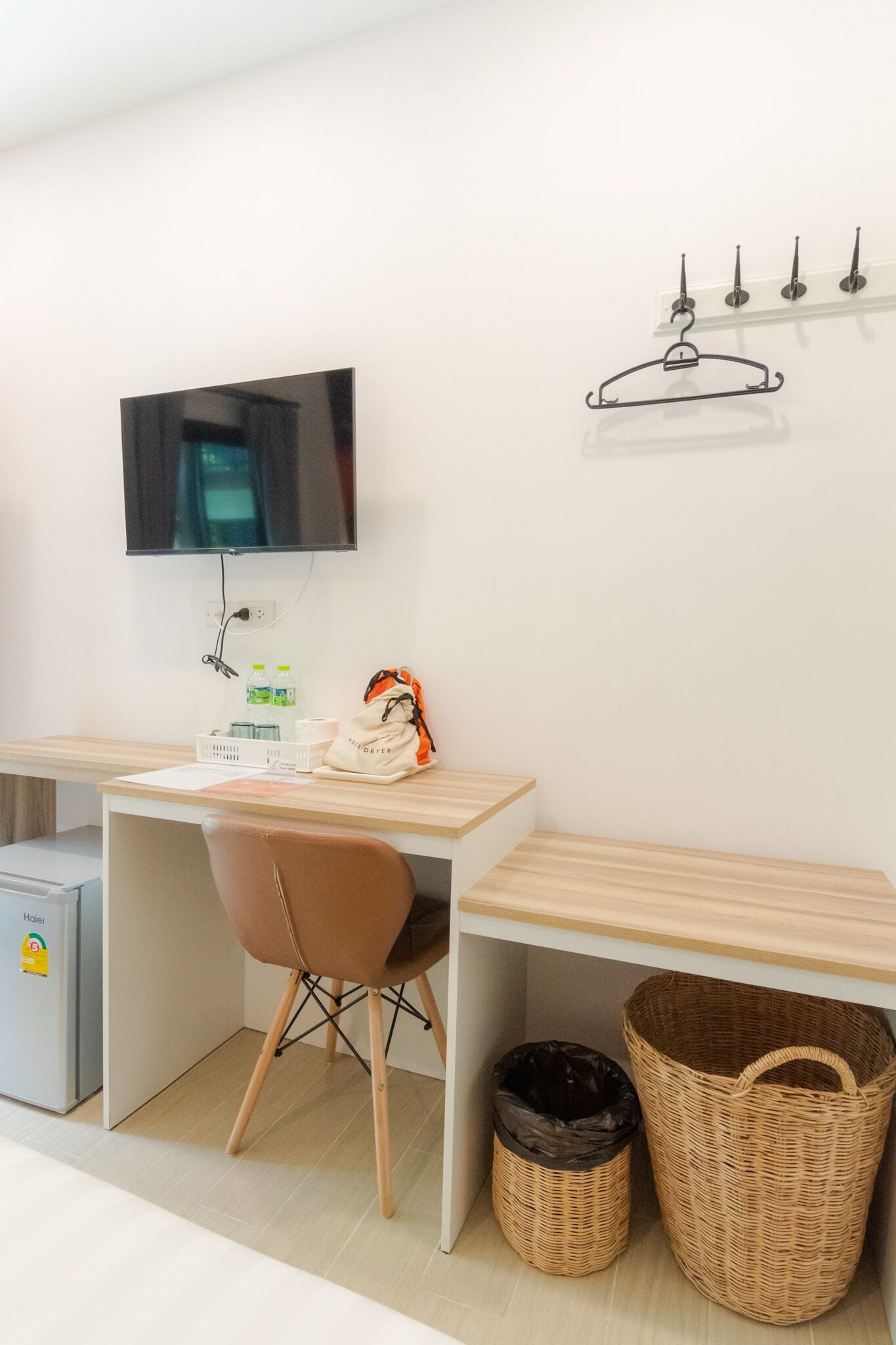 A hotel room with a wooden desk, a chair, a TV mounted on the wall, a coat rack, a trash bin, and a wicker basket.