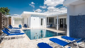 A pool area with blue lounge chairs, a clear pool, and a white building with a tiled wall. - Ayia Napa Holiday Villa Ch (Ayia Napa)