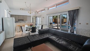 A modern living room with a black leather sectional sofa, a dining area with a table and chairs, and a kitchen in the background. - Ayia Napa Holiday Villa Ch (Ayia Napa)