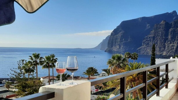 Un balcón con dos copas de vino, con vistas a la costa, palmeras y montañas al fondo.