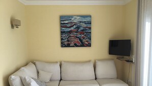 Un salón con un sofá color beige, un reposapiés negro, un cuadro de un paisaje marino, un televisor de pantalla plana y una ventana con cortinas.