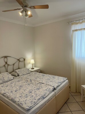 Une chambre à coucher comprenant un lit, une table de chevet, une lampe, un ventilateur de plafond et une fenêtre avec des rideaux.
