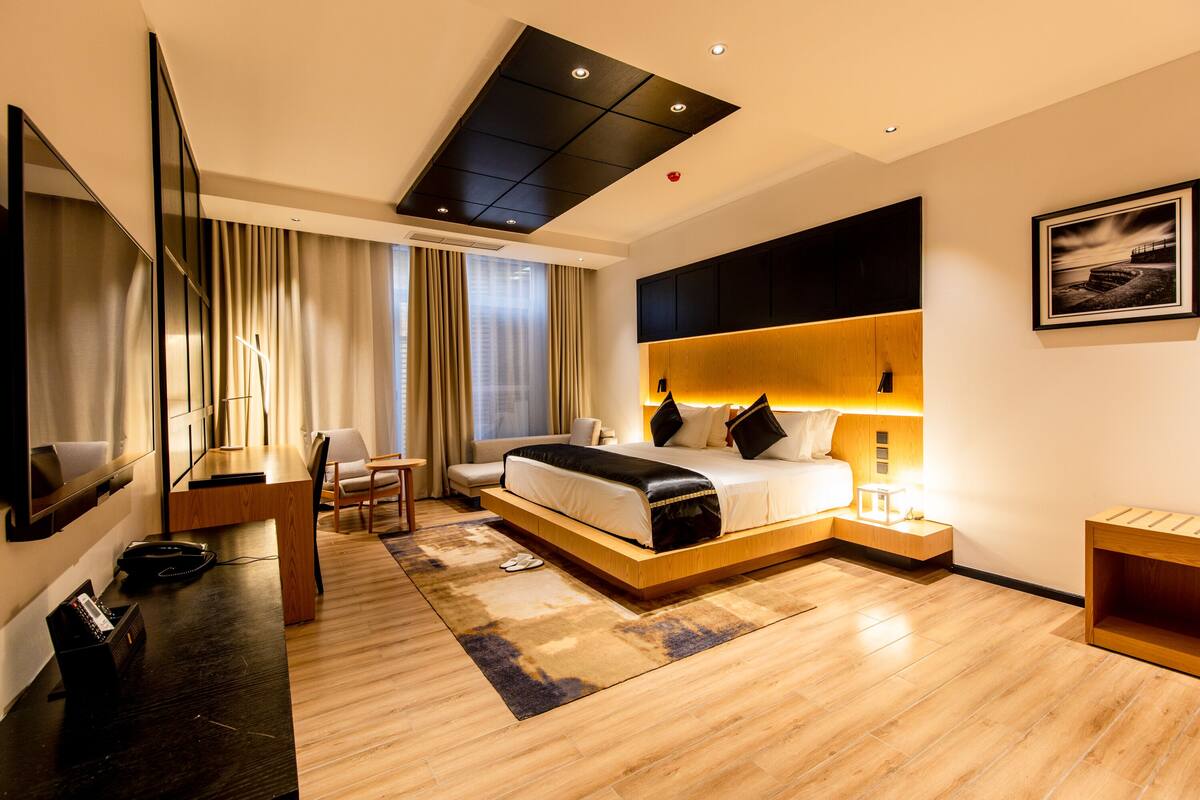 A modern hotel room with a large bed, a desk with a chair, a TV, and a large window with curtains.