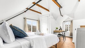 Une chambre avec un plafond en pente, un lit avec une literie blanche, un bureau avec une chaise et une fenêtre avec des rideaux.