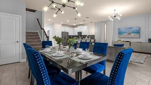 A dining area with a rectangular table set for six, surrounded by blue upholstered chairs with gold studs. There is a modern chandelier with multiple globes above the table.