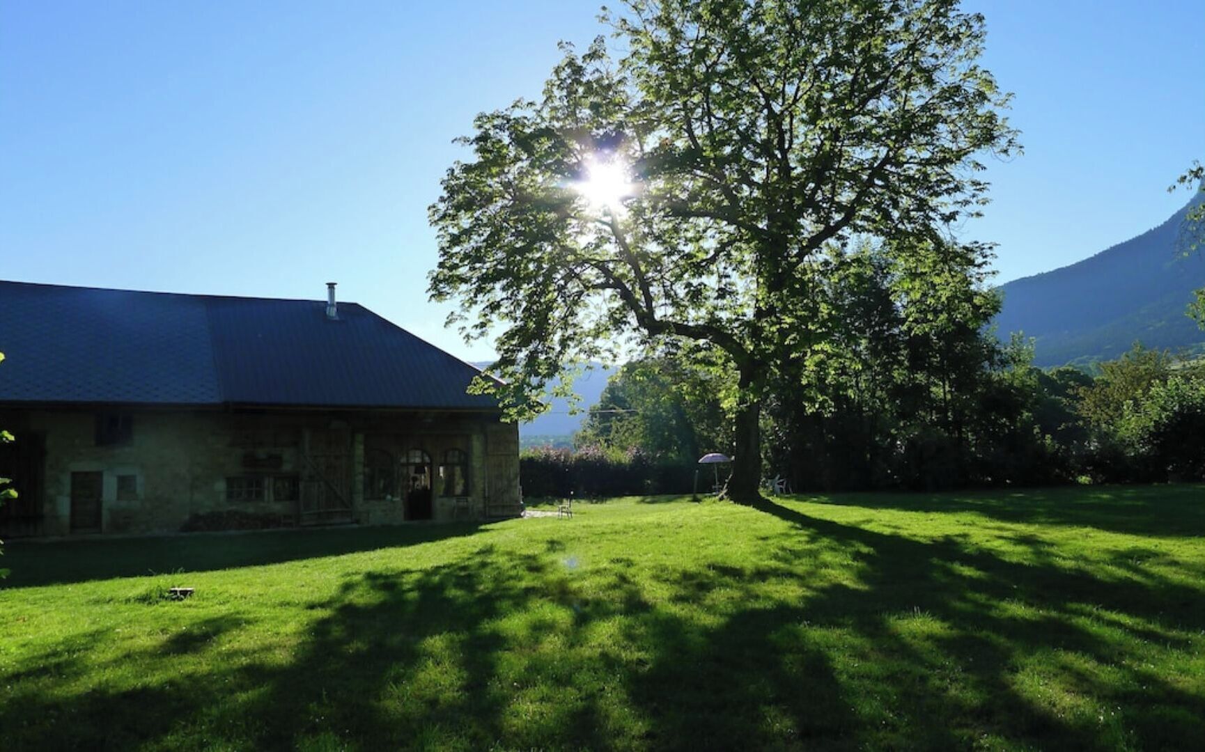 A rustic stone building with a sloped roof, surrounded by a well-maintained lawn and a large tree casting shadows.