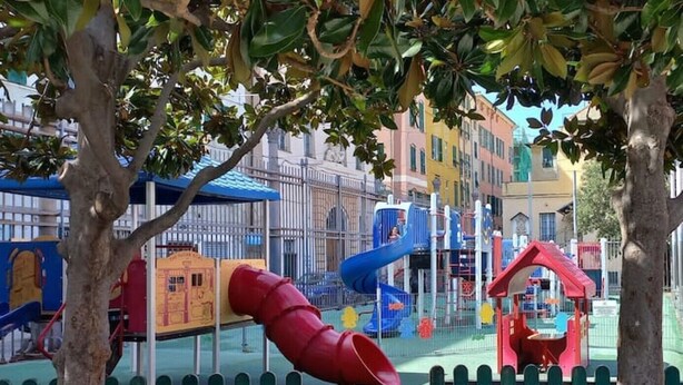 Un parque infantil con toboganes y estructuras para escalar, rodeado por una valla verde y árboles.