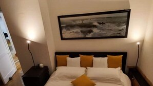 Habitación de hotel con una cama grande, cabecera con varios almohadones, un cuadro de un paisaje marino en la pared y dos mesitas de noche con lámparas.