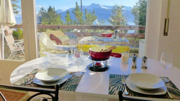 Un tavolo da pranzo con piatti, bicchieri e una pentola rossa, con vista sulle montagne che si intravedono attraverso ampie finestre.