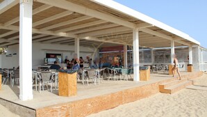An outdoor seating area with tables and chairs, a covered section, and a wooden deck leading to the beach.