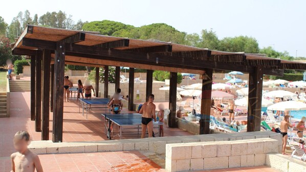 A pool area with a wooden pergola structure, lounge chairs, and a pool table.