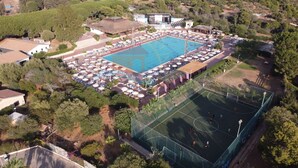 An aerial view of a resort with a large swimming pool, a tennis court, and multiple beach umbrellas.