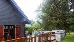 Una casa con terraza de madera, zona de barbacoa de ladrillo y un gran árbol al fondo.