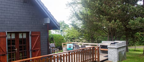 Une maison avec une terrasse en bois, un espace barbecue en briques et un grand arbre en arrière-plan.
