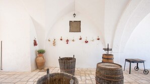 Um interior rústico com soalho de pedra, barris de madeira, uma mesa pequena e um vaso com planta.
