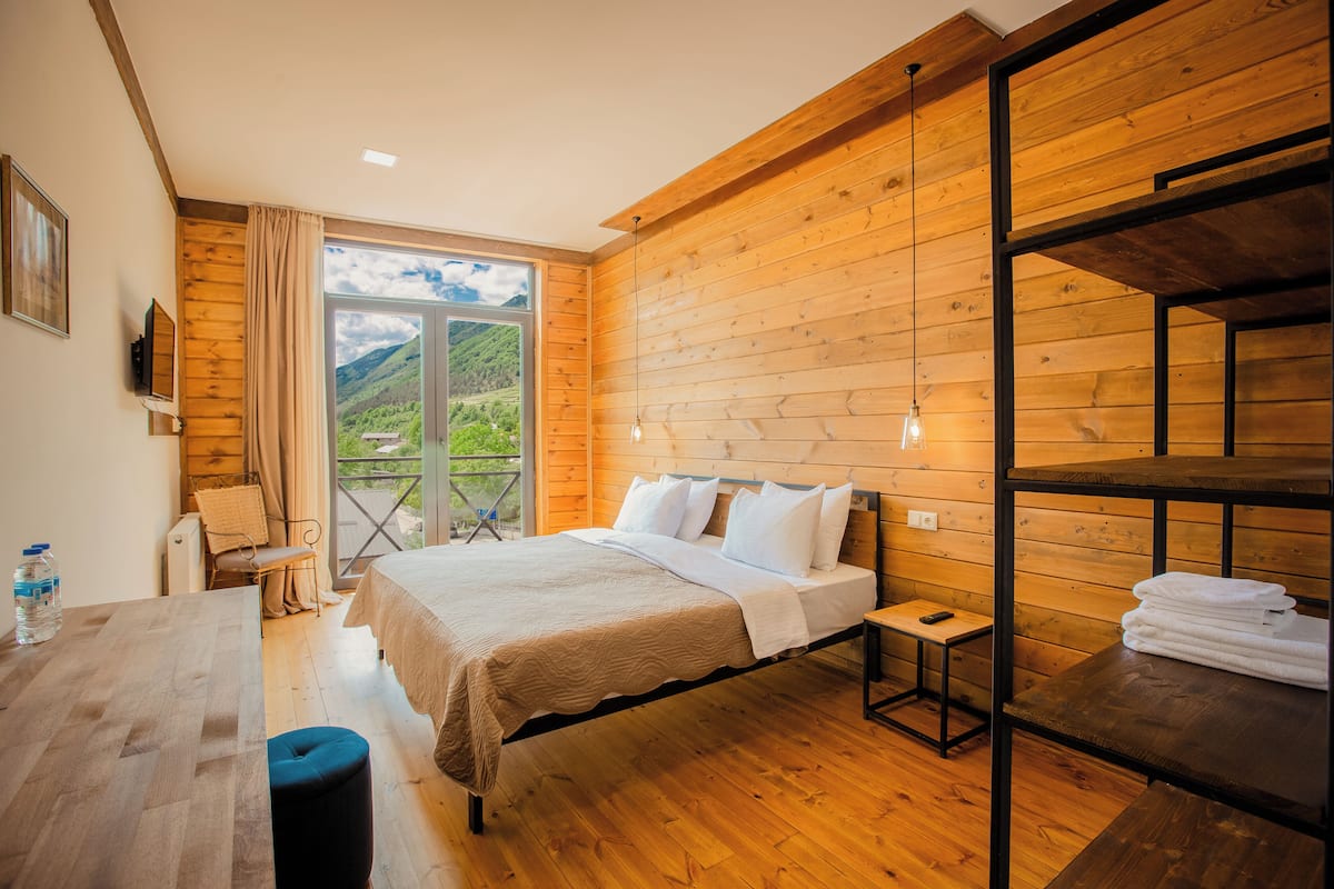 A wooden-paneled bedroom with a bed, a small table, a chair, and a view of greenery outside.